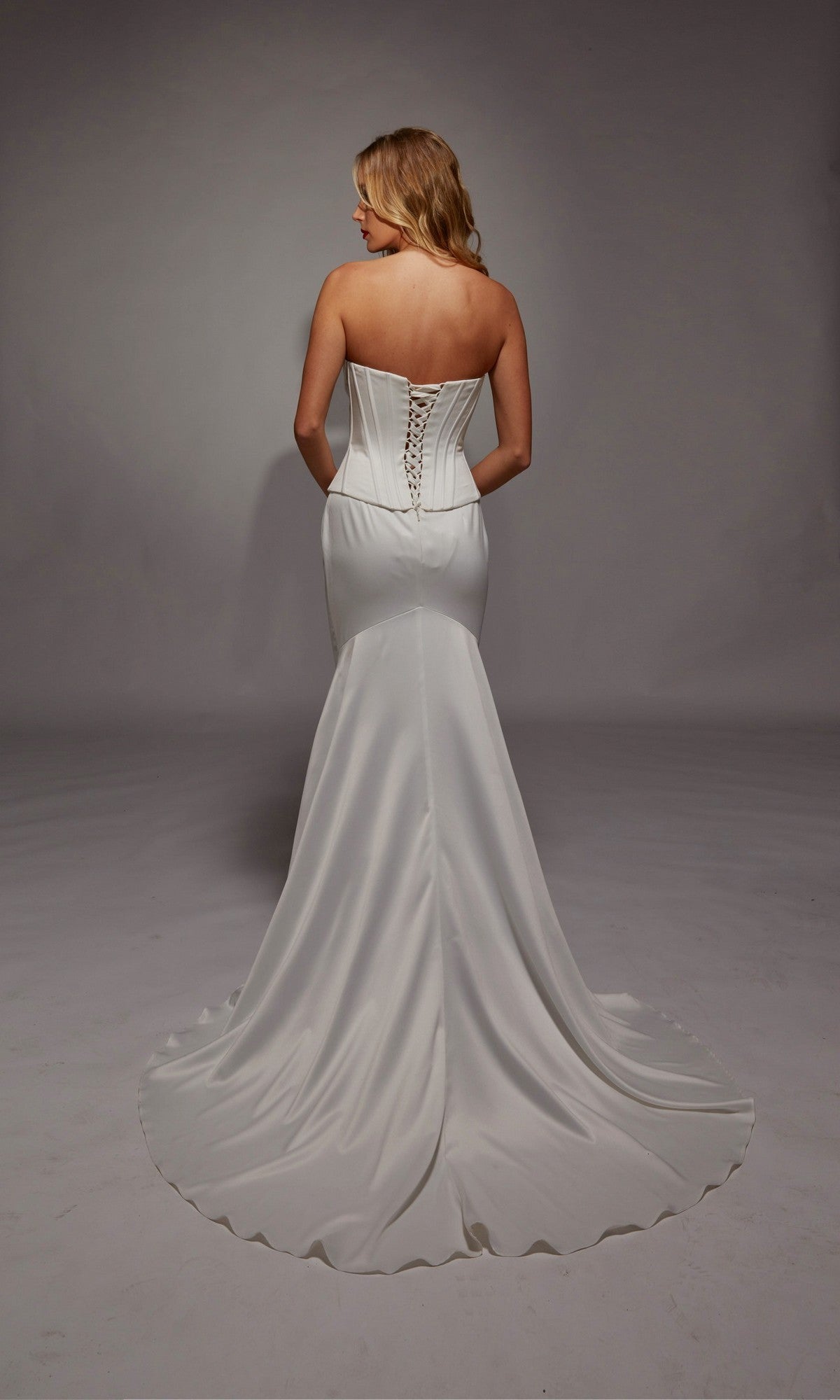 A woman wears the Alyce 7136 long white dress, featuring a strapless satin corset bodice, lace-up back, and sweep train, as she stands with her back to the camera.